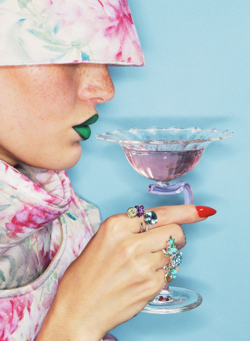 Close-up of a model wearing a floral hat and green lipstick holding a cocktail glass adorned with multiple gemstone rings against blue background.
