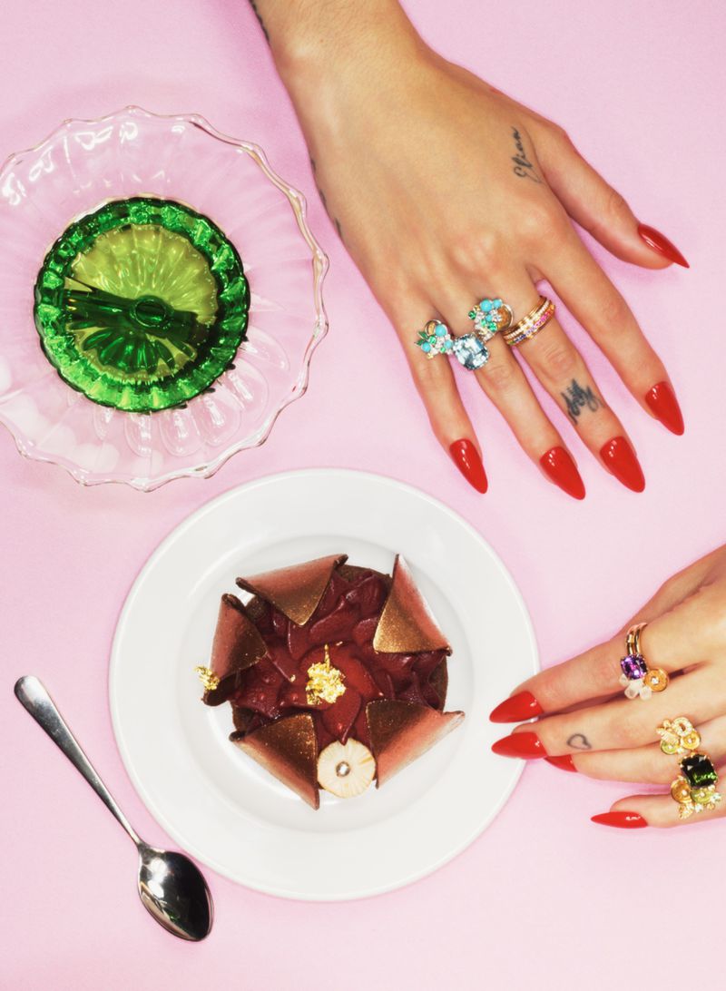 Close-up top-down view of hands adorned with statement rings and long red nails next to a dessert and green gelatin dessert