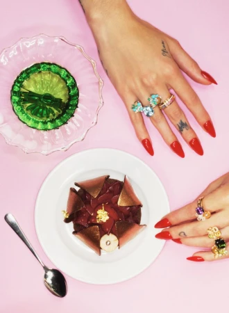 Close-up top-down view of hands adorned with statement rings and long red nails next to a dessert and green gelatin dessert