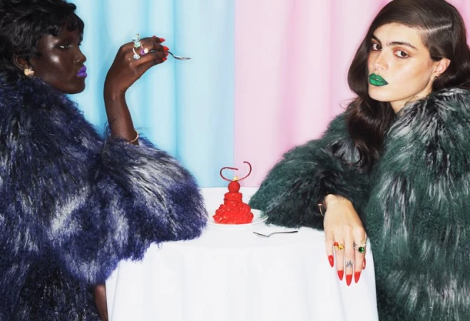 Two models in faux fur coats seated at a table with a red pastry against a blue and pink backdrop for V Magazine.