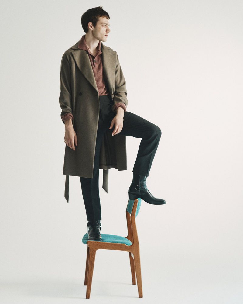 A man wearing a brown coat and dark trousers standing with one foot on a wooden chair against a white background.