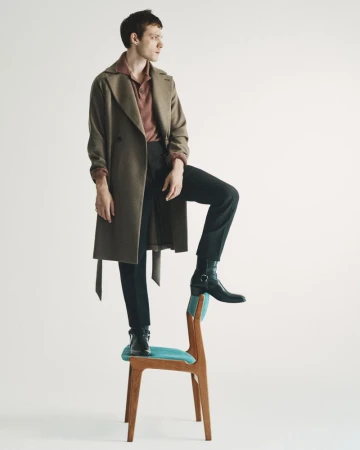 A man wearing a brown coat and dark trousers standing with one foot on a wooden chair against a white background.