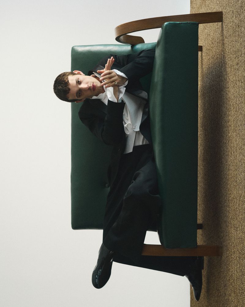 Actor Matteo Oscar Giuggioli seated on a green leather sofa wearing a dark suit and white shirt for Style Magazine Italy.