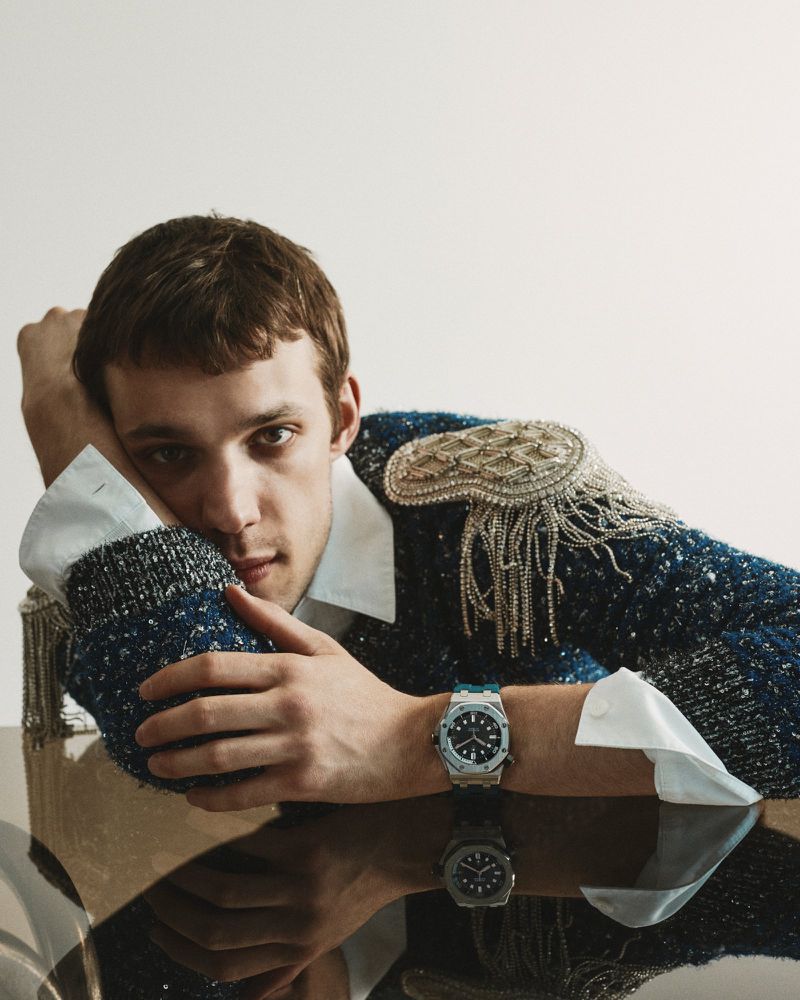Young man resting his head on his hand wearing a blue textured sweater with embellished shoulders and a luxury wristwatch