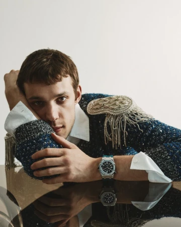 Young man resting his head on his hand wearing a blue textured sweater with embellished shoulders and a luxury wristwatch
