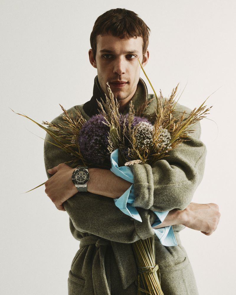 Young man in an olive green coat holding a bouquet of dried flowers and purple alliums against a white background