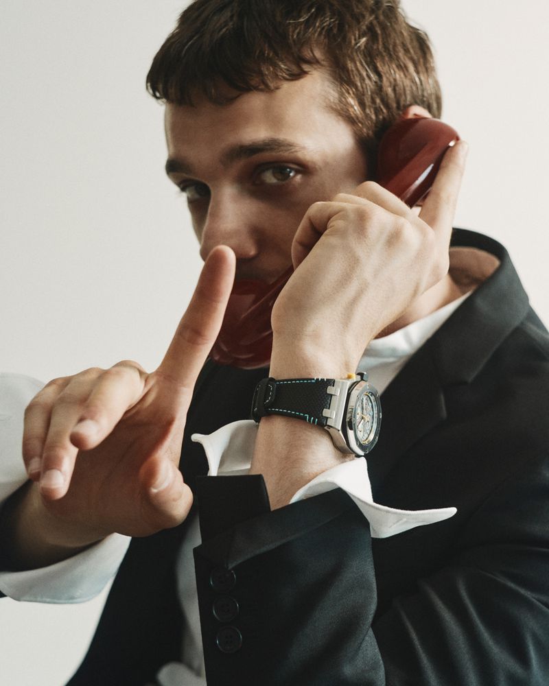 Young man holding a red phone receiver to his ear while gesturing with his index finger, wearing a dark blazer over a white shirt and a wristwatch.