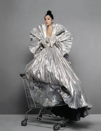 Model standing inside a metal shopping cart wearing an oversized pleated silver gown against a plain grey backdrop.