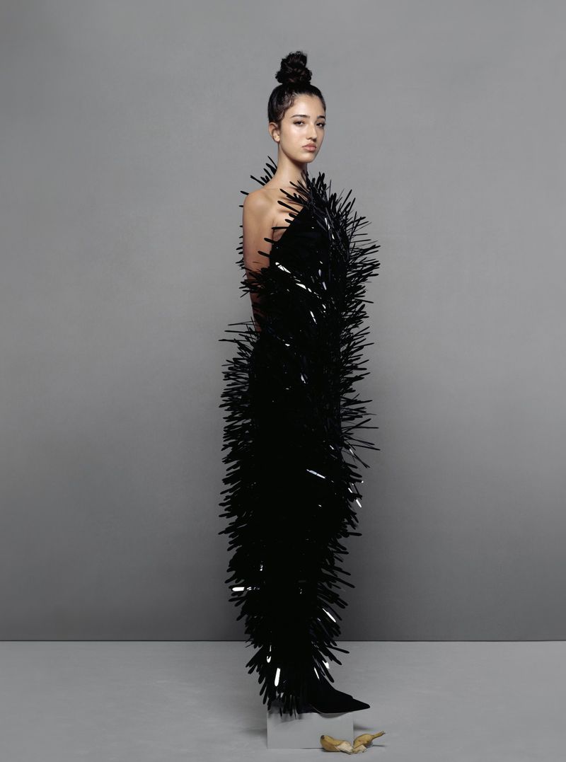 Model in a structured black fringed dress with high top-knot hair against a grey seamless background