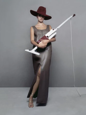 Model wearing a wide-brimmed hat and brown leather dress holding a vacuum cleaner against a grey studio backdrop.