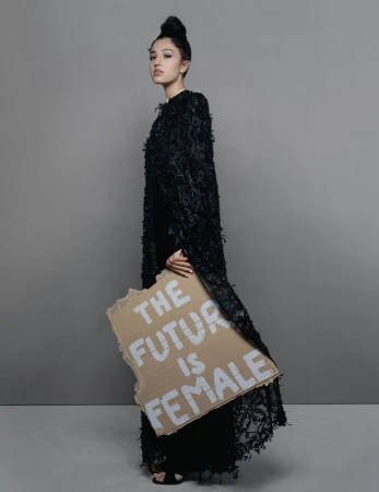 Model in textured black coat holding a cardboard sign against a grey backdrop
