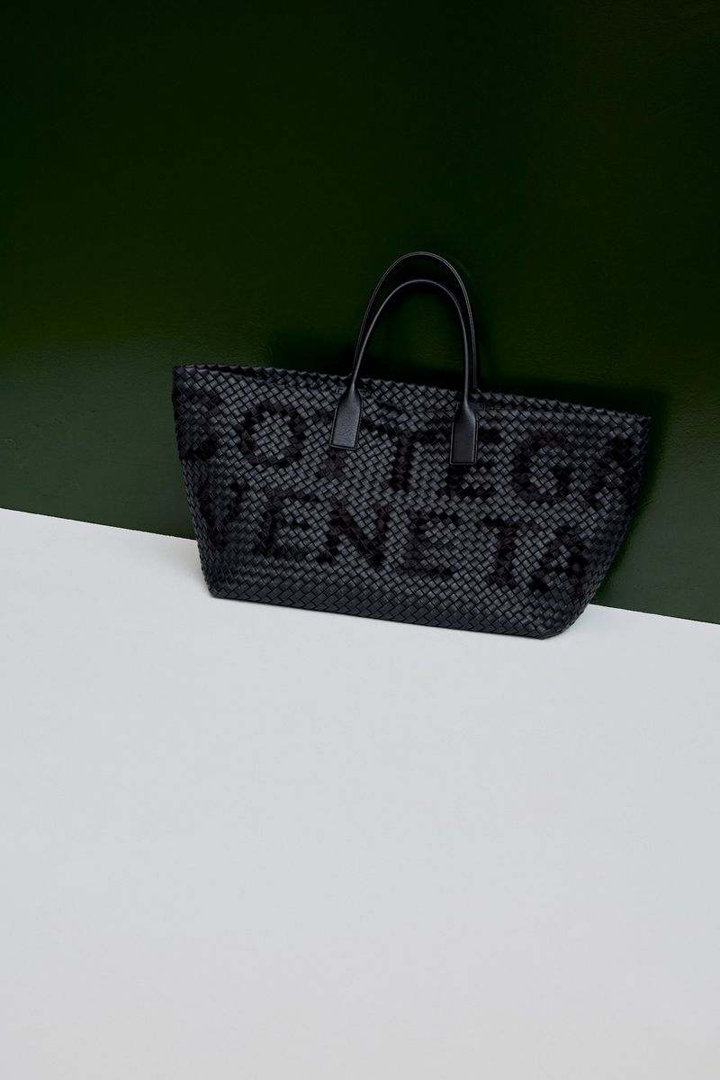 Black woven leather tote bag with top handles resting on a white and dark green studio surface
