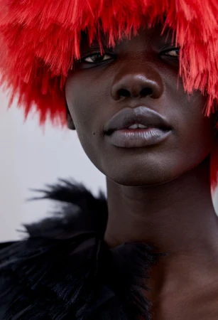 Close-up portrait of a model wearing a vibrant red feathered hat and a black textured garment against a plain white background.