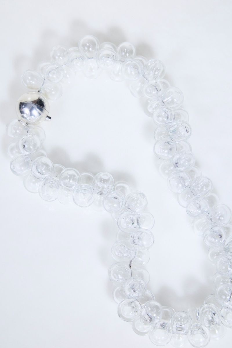 Close-up of a clear glass bubble necklace with a metallic sphere clasp resting against a bright white background.