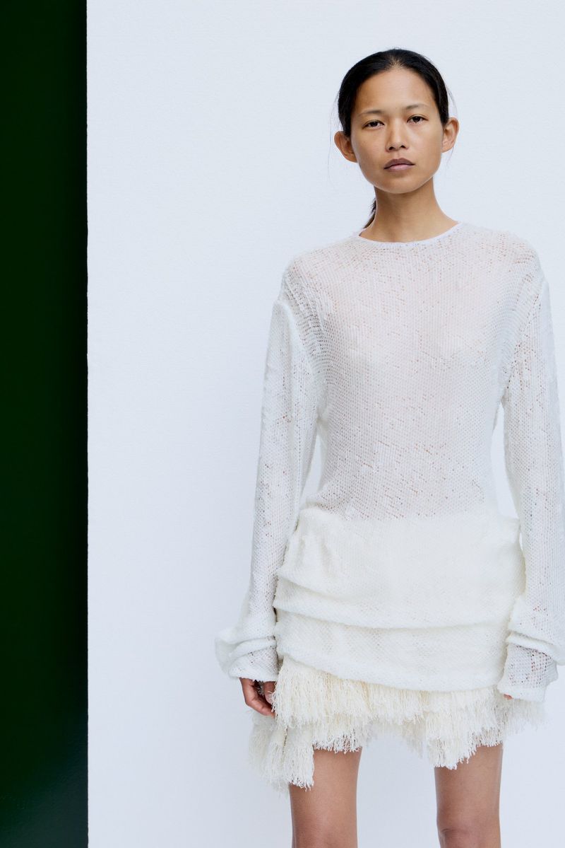 Model wearing a textured white knit top and fringed miniskirt against a pale studio backdrop with a dark vertical stripe
