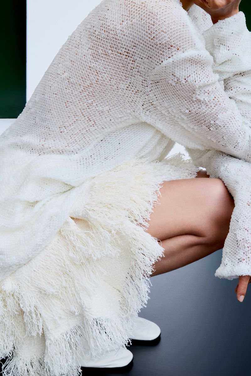 Model crouching wearing a textured white fringe skirt and knit sweater with white boots against a plain background