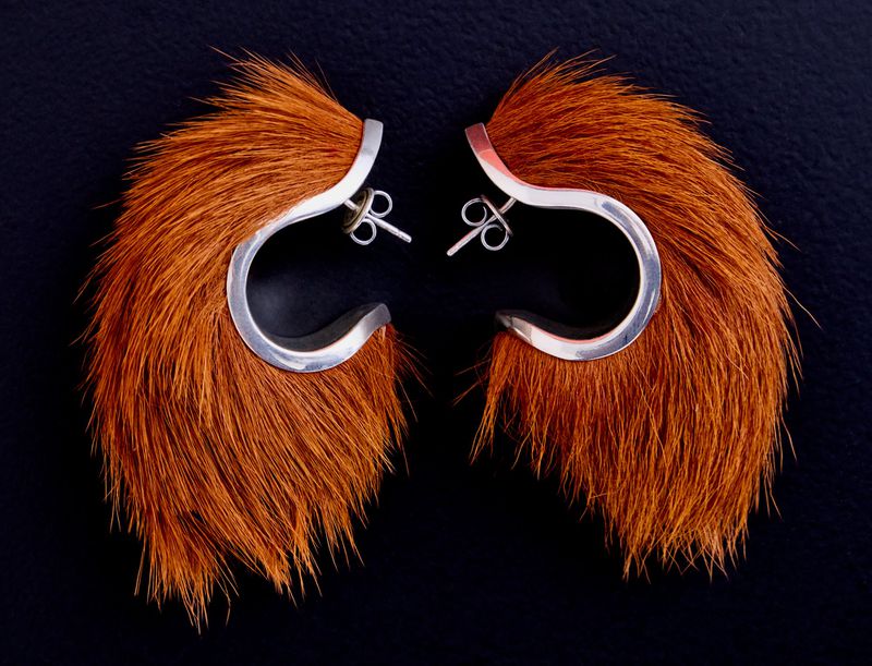 Close-up product shot of a pair of sculptural silver earrings trimmed with long brown animal hair against a dark background