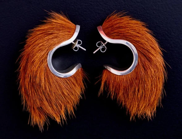 Close-up product shot of a pair of sculptural silver earrings trimmed with long brown animal hair against a dark background
