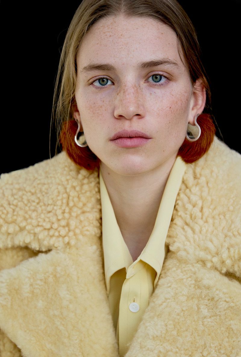 Portrait of a model with freckles wearing a yellow textured coat and pale yellow shirt against a black background.