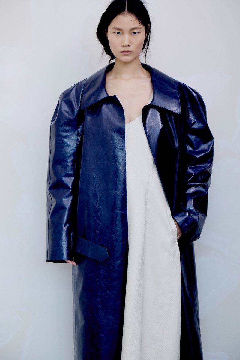 Model wearing a glossy blue leather coat and white dress against a plain white studio background