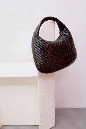 Dark brown leather woven intrecciato handbag displayed on a white sculptural pedestal in a minimal studio setting.