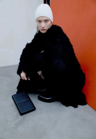 Model in a black faux fur coat and white knit beanie crouching next to a black quilted handbag