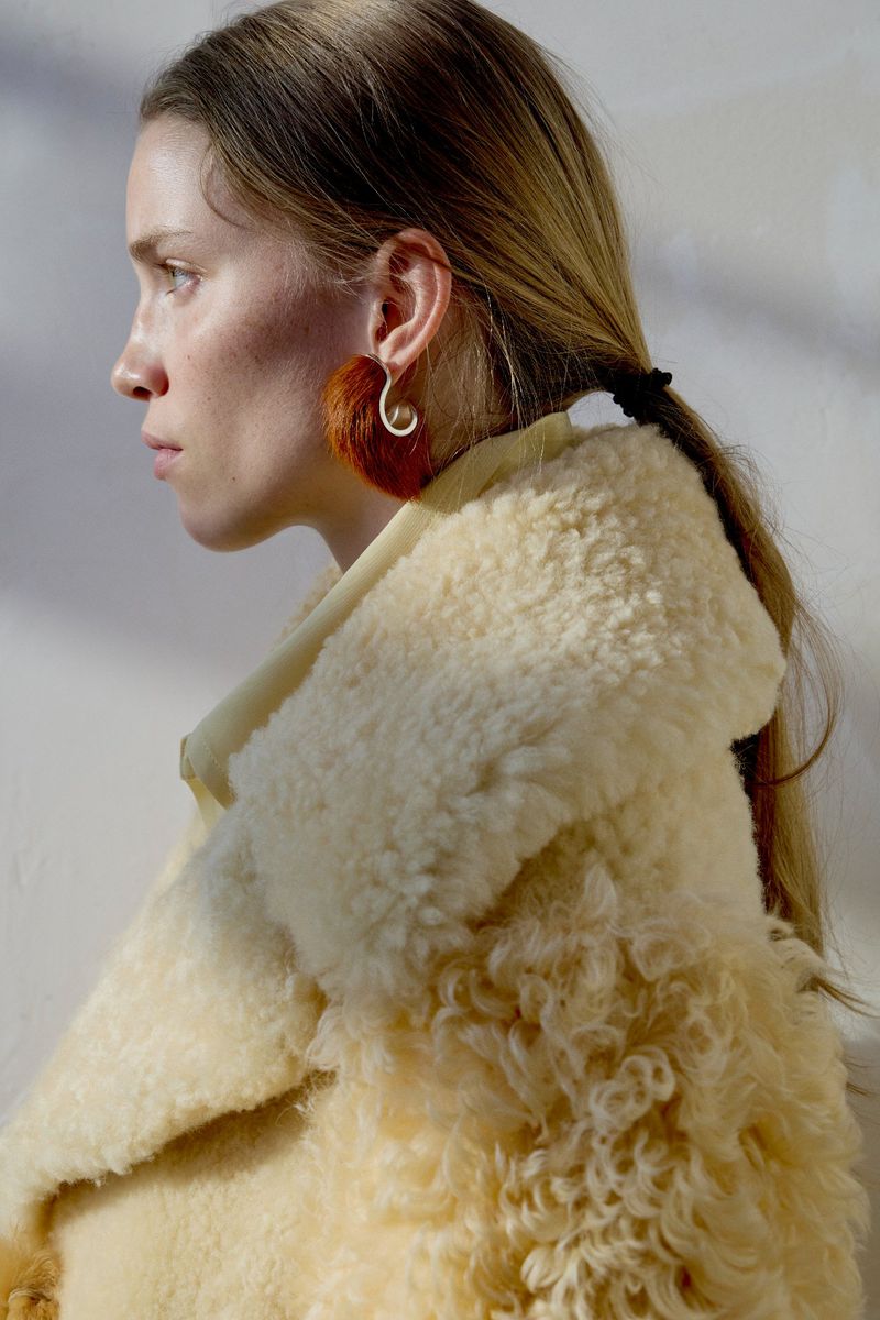 Profile portrait of a woman wearing a cream shearling coat and a distinctive reddish-brown earring against a plain background