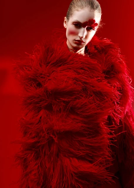 Portrait of a woman in a voluminous red, feathered coat, dramatic red makeup on her face and lips, against a solid red background.