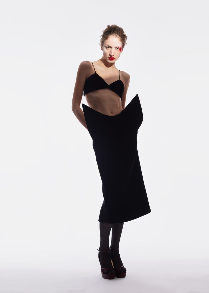 Fashion model poses in a black bralette and skirt against a white backdrop. Red makeup detail on her face, black hose, and platform shoes.