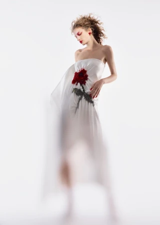 A woman in a strapless white gown with a red rose design poses against a blurred white background in a fashion studio setting.