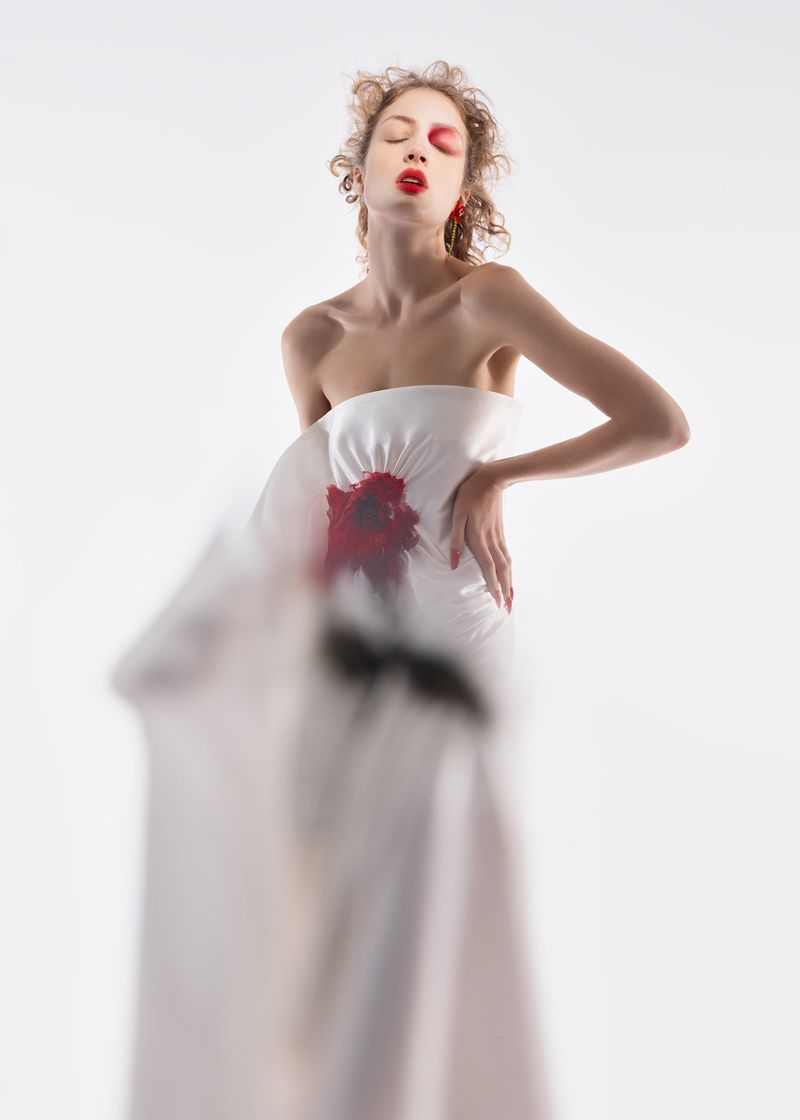 Elegant woman in white strapless dress with artistic red and black flower design posing in a bright photo studio, fashion-forward.