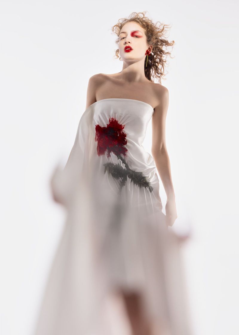 A young woman poses in a flowing white dress with a painted rose design against a stark white backdrop in a fashion-style portrait.