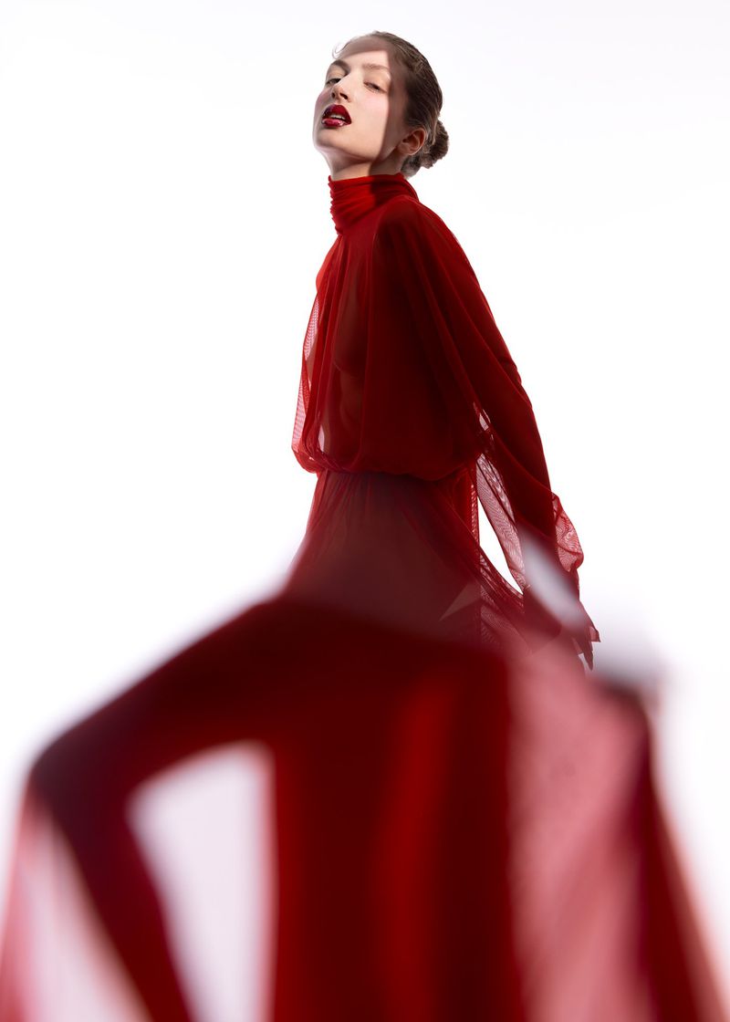 Young woman with red lipstick wears sheer red turtleneck dress against a white background in a studio fashion portrait.