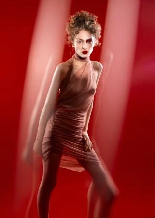 Model with a curly updo in a sheer dusty rose ruched halter dress against a vibrant red backdrop with motion streaks.