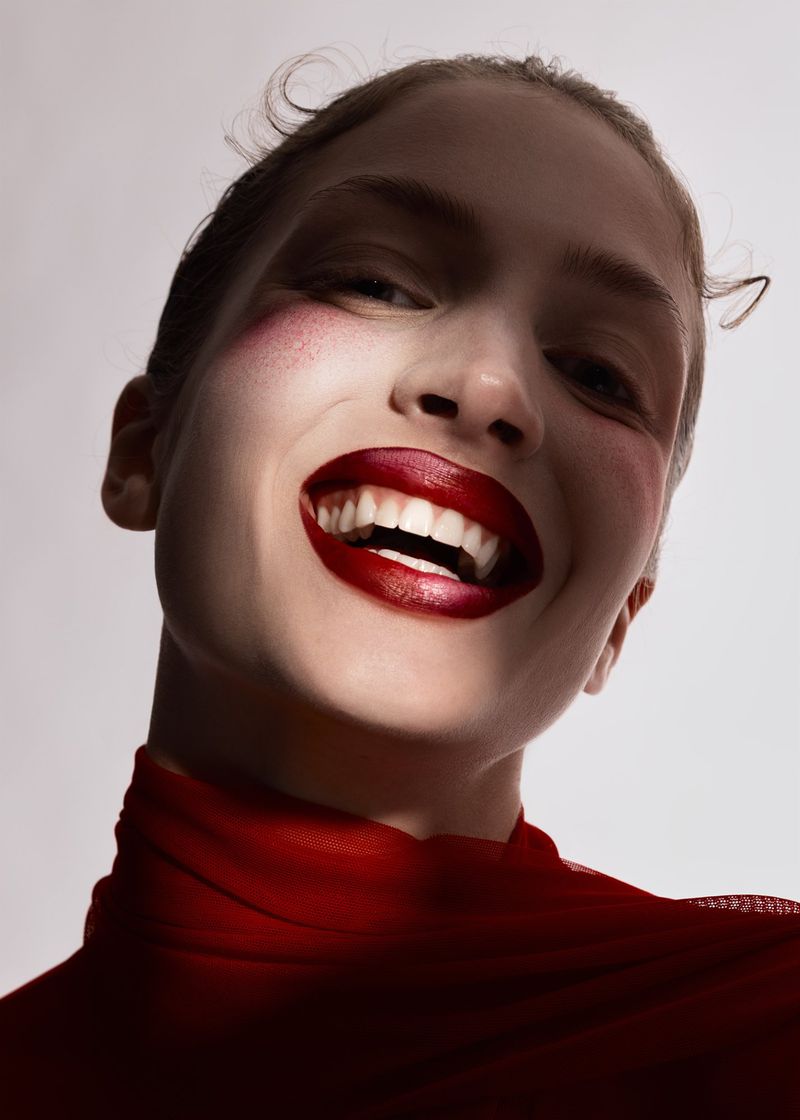 Close-up beauty shot of a smiling young adult woman with dark red lipstick and blush, against a neutral backdrop and soft lighting.