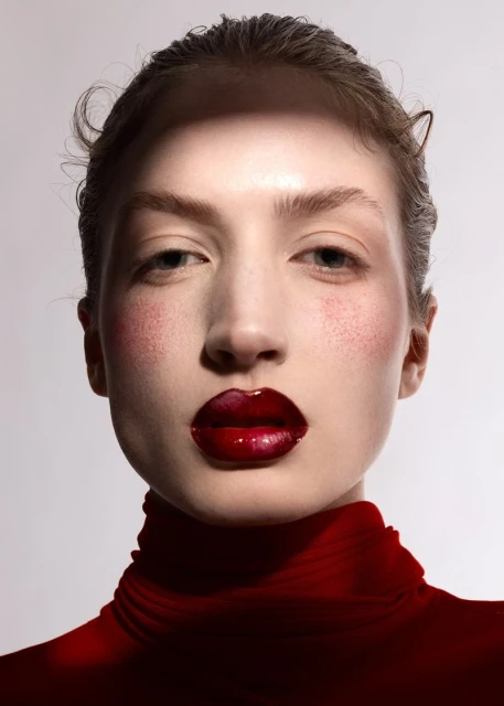 Close-up beauty shot of a woman with glossy red lips and red blush, wearing a red turtleneck against a light neutral background.