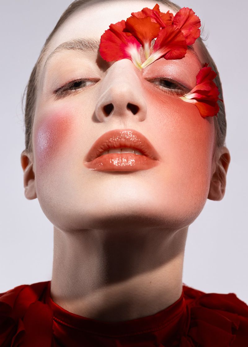 Close-up portrait of a fair-skinned adult female model wearing red lipstick and flower petals on her face against a grey background.