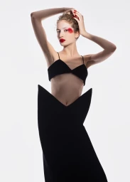 Model posing with hands on head wearing a black bralette, structural pointed skirt, red lipstick, and red eye makeup on white backdrop.