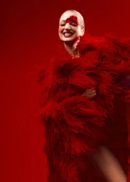 Model wearing a vibrant red feathered coat with flower petal eye makeup against a solid red background