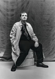 Monochrome portrait of French actor Karim Leklou sitting on crates wearing a trench coat, dark shirt, trousers, and patterned scarf.