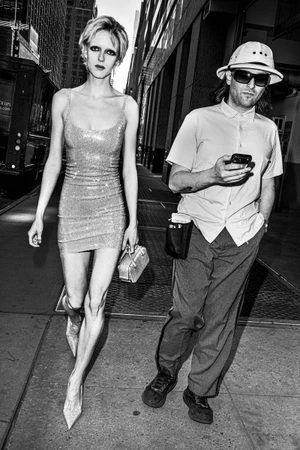 Black and white street photograph of a blonde model walking alongside a man looking at a phone in the city