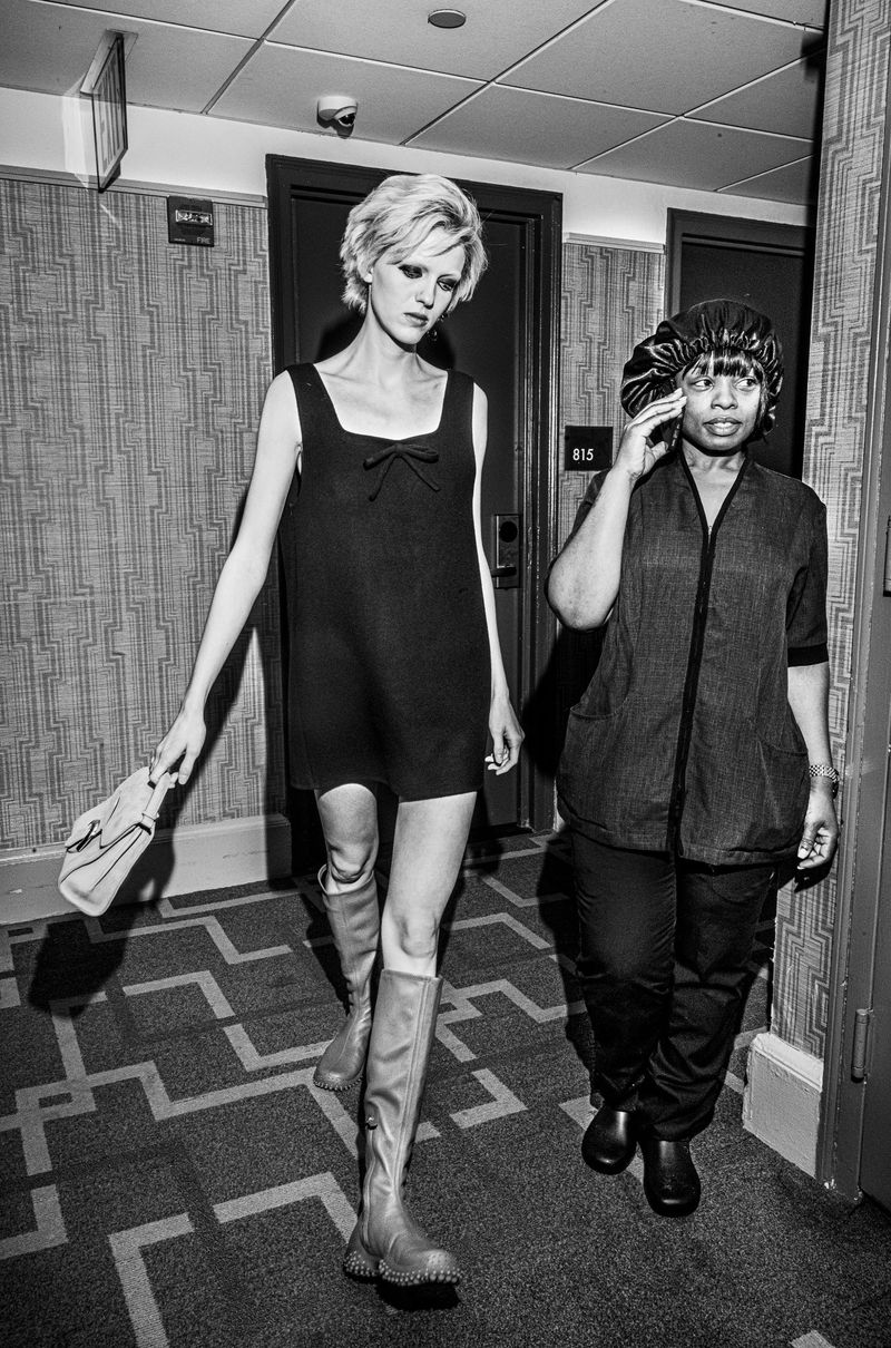 Black and white editorial photo of a model in a short dark dress walking down a hotel hallway alongside a hotel staff member.
