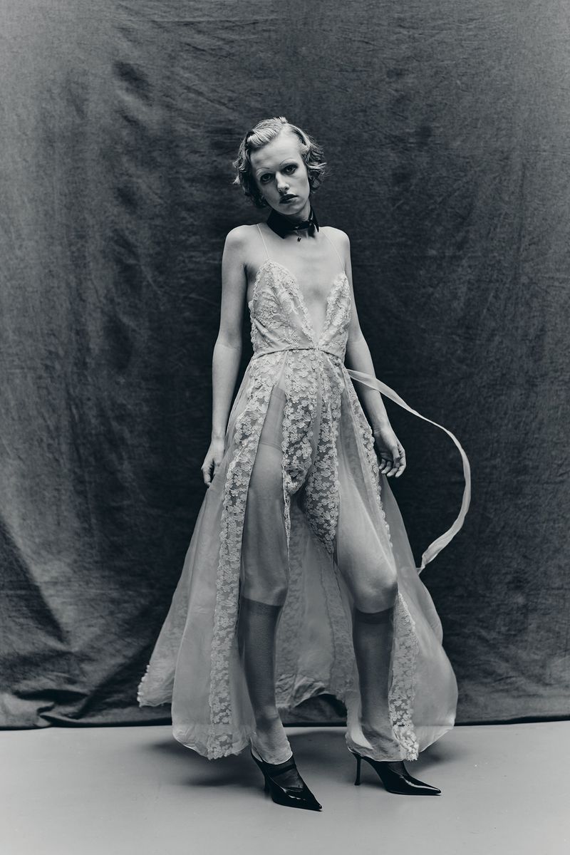 Model Sasha Krivko wearing a sheer lace dress with a high slit against a textured grey backdrop for Crash Magazine