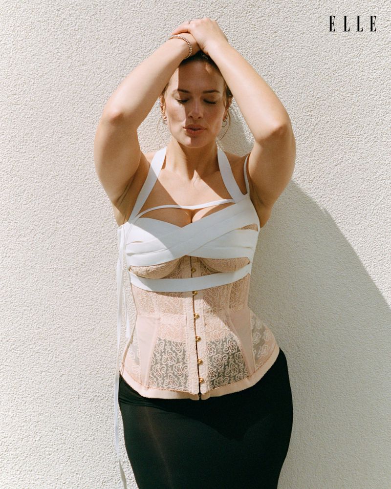 Ashley Graham wearing a structured patterned corset and dark skirt with hands on head against a textured white wall.