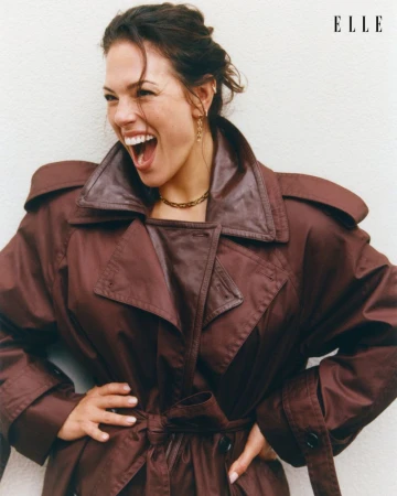 Ashley Graham laughing while wearing a brown leather trench coat against a white background for Elle Netherlands.