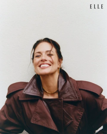 Ashley Graham smiling in a dark brown leather trench coat against a plain white textured background for Elle Netherlands.
