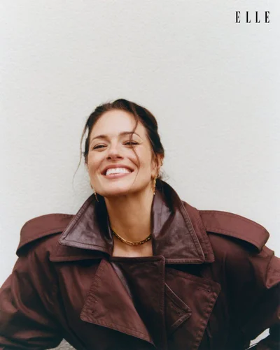 Ashley Graham smiling in a dark brown leather trench coat against a plain white textured background for Elle Netherlands.