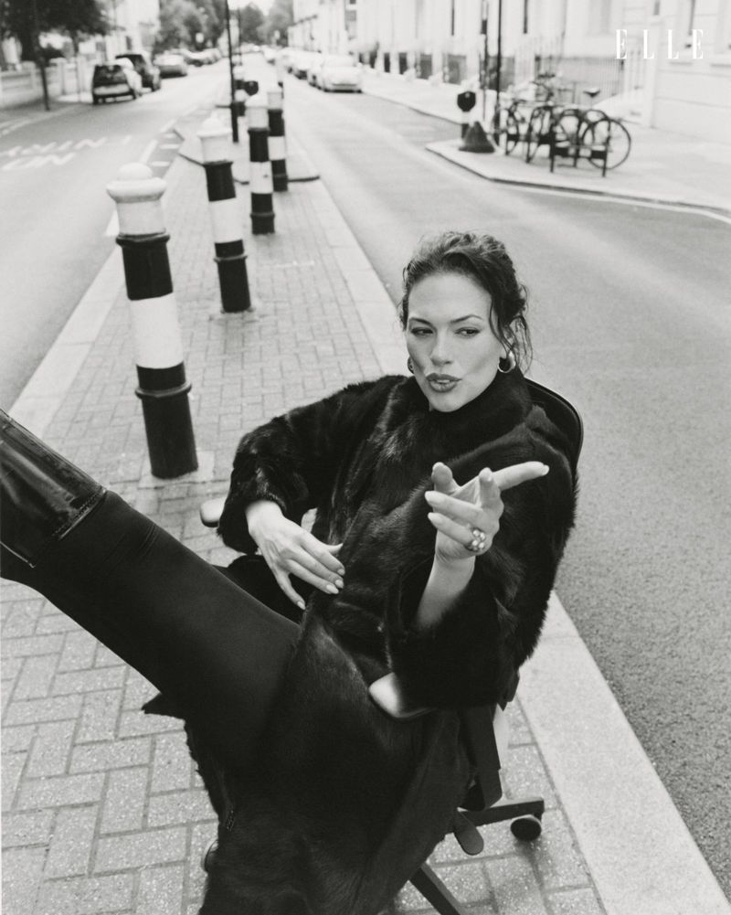 Model Ashley Graham sitting in an office chair on a city street in a black coat with boots raised toward camera
