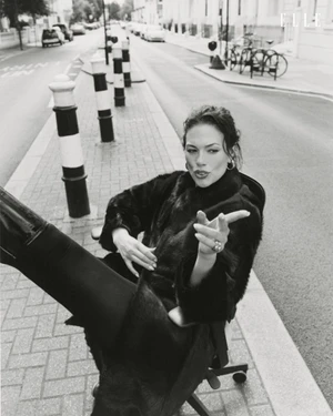 Model Ashley Graham sitting in an office chair on a city street in a black coat with boots raised toward camera