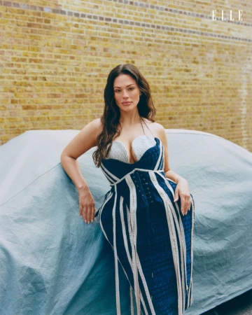 Ashley Graham posing in a structured blue and white dress leaning against a covered car in front of a brick wall
